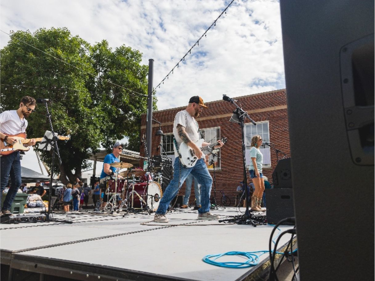 A band performing on an outdoor stage at Brews & Cruise, and in the background, a brick building, a tall tree, and groups of people.